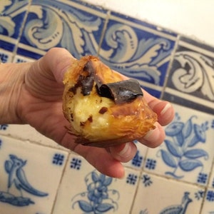 May include: A hand holding a Portuguese custard tart with a flaky pastry crust and a layer of caramelized sugar on top. The tart is being held in front of a tiled wall with blue and white floral designs.