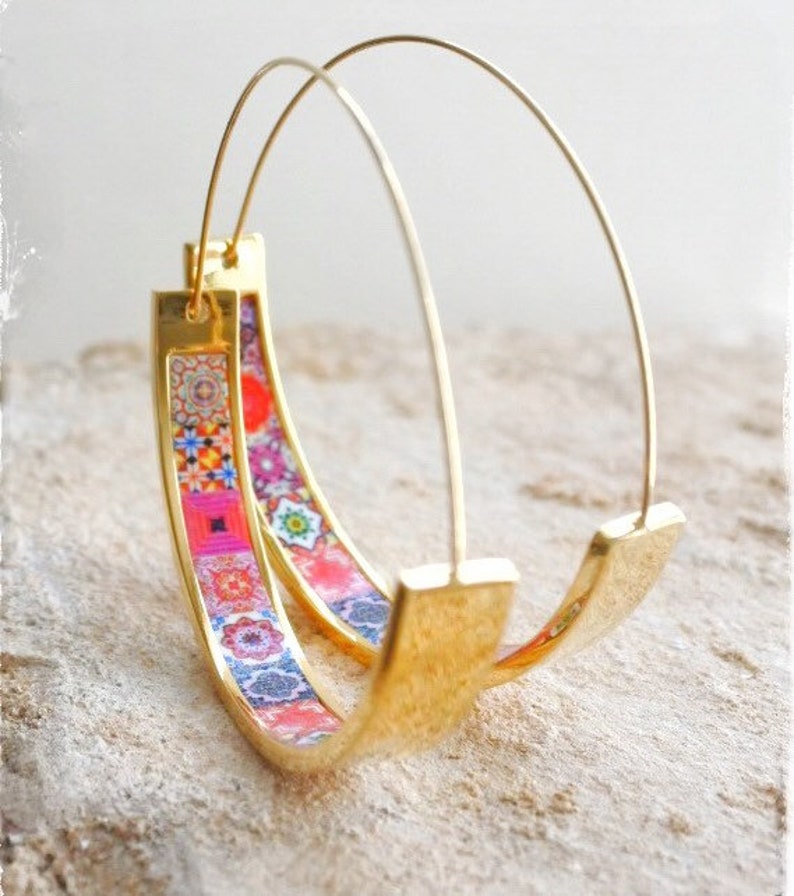 May include: Gold hoop earrings with a colorful patterned design. The pattern features geometric shapes and floral motifs in shades of pink, red, blue, and green.