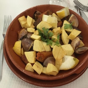 May include: A terracotta bowl filled with a dish of cubed fried potatoes, dark brown meat chunks, and whole clams, garnished with fresh green cilantro leaves and wedges of yellow lemon, served on a matching terracotta plate.