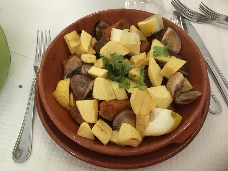 May include: A terracotta bowl filled with a dish of cubed fried potatoes, dark brown meat chunks, and whole clams, garnished with fresh green cilantro leaves and wedges of yellow lemon, served on a matching terracotta plate.