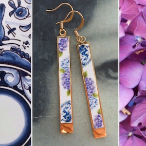 May include: A pair of long, gold dangle earrings with white porcelain rectangular panels featuring a floral pattern in shades of blue and purple. The panels are framed with a gold border.