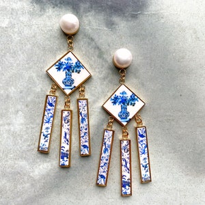 May include: A pair of blue and white porcelain earrings with a dangling design. Each earring features a square tile with a floral pattern and a vase, and four rectangular tiles with a bird and floral pattern.
