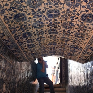 May include: A person taking a photo of a tiled ceiling with a blue and white floral pattern. The ceiling is arched and the person is standing on a set of stairs.