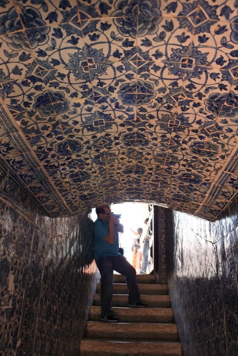 May include: A person taking a photo of a tiled ceiling with a blue and white floral pattern. The ceiling is arched and the person is standing on a set of stairs.