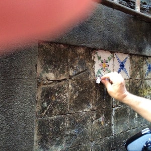 Puede incluir: Primer plano de una pared con azulejos agrietados y desgastados. Los azulejos est&aacute;n decorados con motivos florales y geom&eacute;tricos en tonos azules, blancos y amarillos. Una mano est&aacute; despegando un trozo de papel de uno de los azulejos.