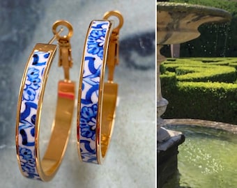 May include: A pair of gold hoop earrings with a white and blue floral pattern. The earrings are about 2 inches in diameter.