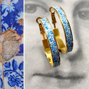 May include: A pair of gold hoop earrings with a white and blue floral pattern. The earrings are approximately 5 cm in diameter.