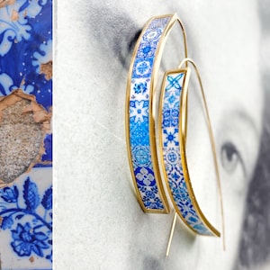 May include: A pair of gold hoop earrings with blue and white patterned designs. The earrings are made of metal and have a simple, elegant design.