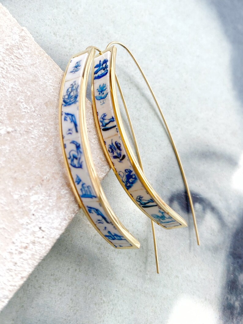 Op de afbeelding: Een paar gouden creolen met een blauw en wit patroon. Het patroon bevat verschillende kleine afbeeldingen, waaronder bloemen, vogels en geometrische vormen.