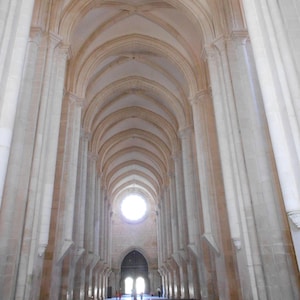 May include: A long, narrow, white-walled hallway with a vaulted ceiling and rows of wooden pews. A large, round window at the end of the hallway lets in light.
