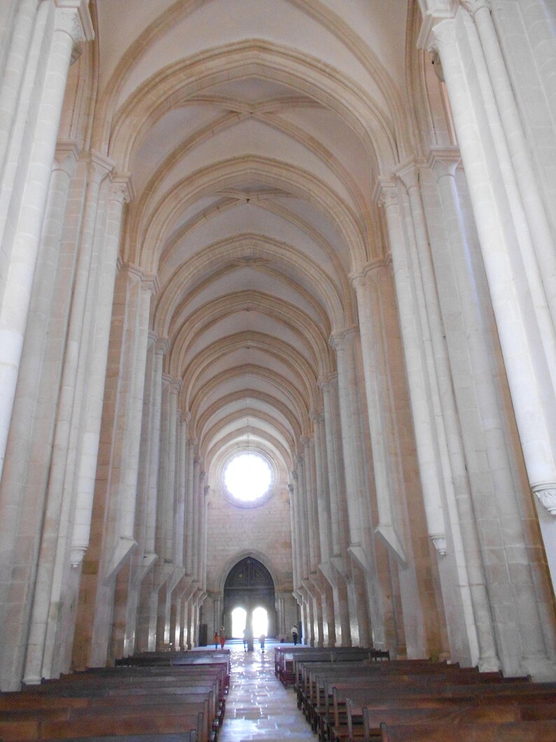 May include: A long, narrow, white-walled hallway with a vaulted ceiling and rows of wooden pews. A large, round window at the end of the hallway lets in light.