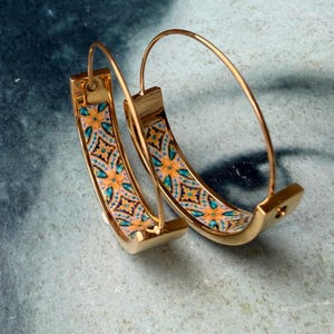 May include: A pair of gold-colored hoop earrings. The earrings have a rectangular, curved design with a colorful pattern of orange, teal, and black geometric shapes. The hoops are set against a muted blue background.