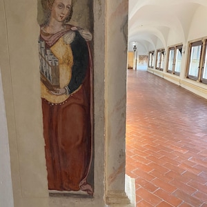 May include: A vertical wall fresco depicting a figure in a yellow bodice and dark red skirt holding a small pipe organ. The painting is situated on a pillar next to a long hallway with a terracotta tiled floor and a series of arched windows.