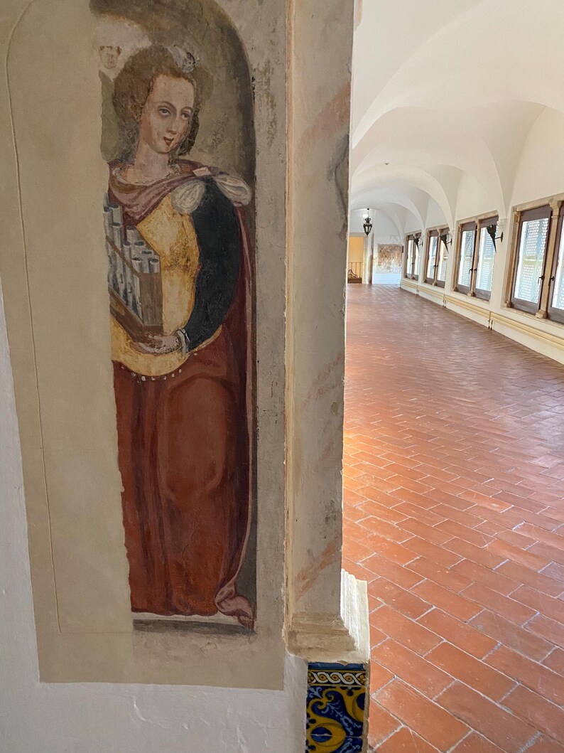May include: A vertical wall fresco depicting a figure in a yellow bodice and dark red skirt holding a small pipe organ. The painting is situated on a pillar next to a long hallway with a terracotta tiled floor and a series of arched windows.