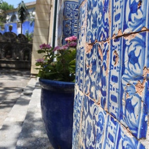May include: A close-up of a blue and white tiled wall with intricate floral patterns. The tiles are cracked and chipped, showing the red clay underneath. A blue planter with green plants sits in front of the wall.