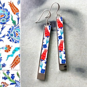 May include: A pair of silver dangle earrings with a white background and red, blue, and green floral designs. The earrings are inspired by Turkish tile patterns.