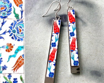 May include: A pair of silver dangle earrings with a white background and red, blue, and green floral designs. The earrings are inspired by Turkish tile patterns.