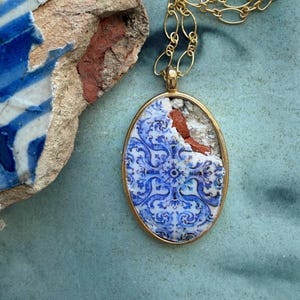 May include: Gold-tone necklace with an oval pendant. The pendant features a fragment of a blue and white patterned tile, set in a gold frame. The tile has a floral design. The necklace is on a teal surface.