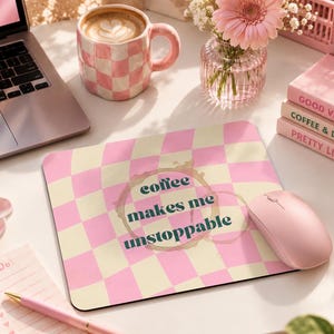 Könnte beinhalten: Ein rechteckiges Mauspad mit einem rosa-cremefarbenen Karomuster und einer Kaffeering-Grafik in der Mitte. Der Text "coffee makes me unstoppable" ist in dunkelgrünen Buchstaben über dem Kaffeering-Design aufgedruckt.