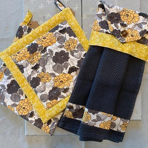 May include: A quilted pot holder and hanging kitchen towel set. The pot holder is square with a yellow border and floral pattern in yellow, gray, and white. The towel is dark gray with a floral pattern and yellow accents.
