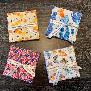 May include: Four square fabric coasters with various patterns, including bees, cacti, butterflies, and an American flag design. Each set is tied with a white ribbon printed with "Sunshine Home" and "by kedesigns".