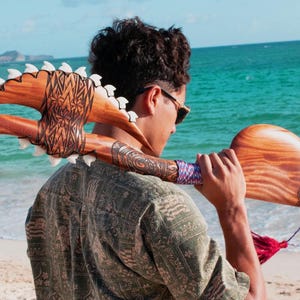 May include: A person holding a wooden carved weapon with a shark tooth design. The weapon is brown and white with intricate carvings. The person is standing on a beach with the sea in the background.