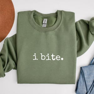 May include: An olive green sweatshirt with the phrase "i bite." printed in white. The long-sleeved top is made of a soft material and is suitable for casual wear. The sweatshirt is folded, with a brown hat and blue jeans in the background.