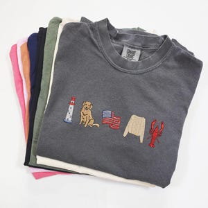 May include: A stack of Comfort Colors sweatshirts in various colors, including pink, navy, and green. The dark gray sweatshirt on top features embroidered images of a lighthouse, dog, American flag, sweater, and lobster.
