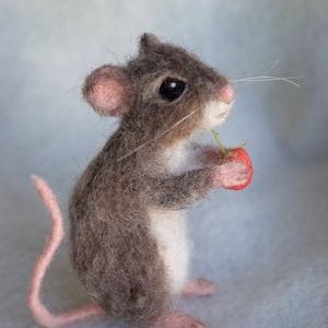 Puede incluir: Un pequeño ratón de fieltro de aguja, gris y blanco, sosteniendo una fresa roja. El ratón tiene una cola rosa y está de pie sobre una superficie gris claro.