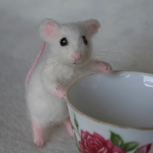 Puede incluir: Un ratón blanco de fieltro con orejas rosas y cola rosa, mirando una taza de té con un diseño de rosa rosa.