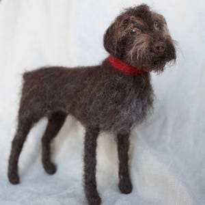May include: A brown and white needle felted dog with a red collar. The dog is standing on a white surface.