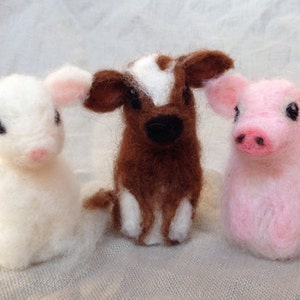 Pode incluir: Tr&ecirc;s figuras de animais de fazenda feitas &agrave; m&atilde;o em feltro: um cordeiro branco, uma vaca marrom e branca e um porco rosa. Cada um tem pequenos olhos pretos e detalhes em rosa nas orelhas e focinho. As figuras est&atilde;o em um fundo neutro.