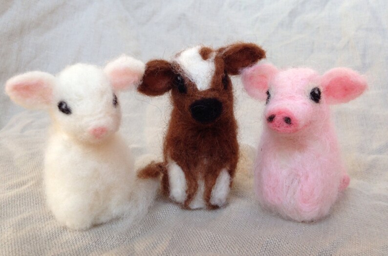 Pode incluir: Tr&ecirc;s figuras de animais de fazenda feitas &agrave; m&atilde;o em feltro: um cordeiro branco, uma vaca marrom e branca e um porco rosa. Cada um tem pequenos olhos pretos e detalhes em rosa nas orelhas e focinho. As figuras est&atilde;o em um fundo neutro.