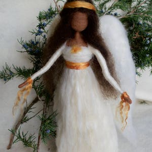 May include: A white felt angel with brown hair, a gold crown, and gold accents on the dress. The angel has white wings and is standing on a white surface.