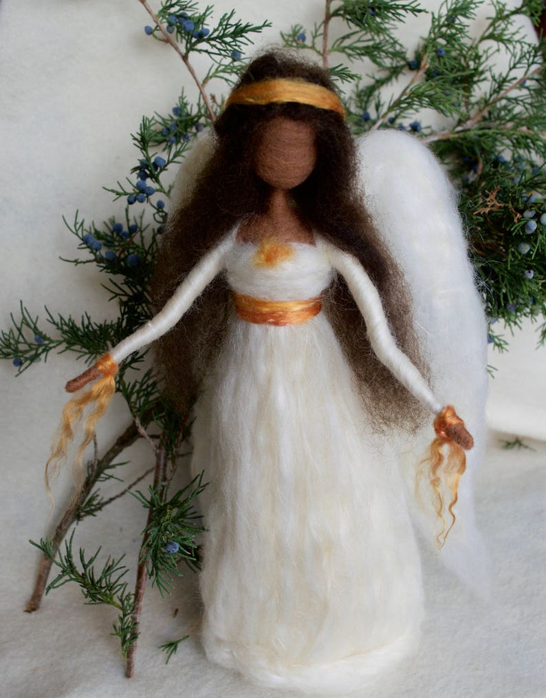 May include: A white felt angel with brown hair, a gold crown, and gold accents on the dress. The angel has white wings and is standing on a white surface.