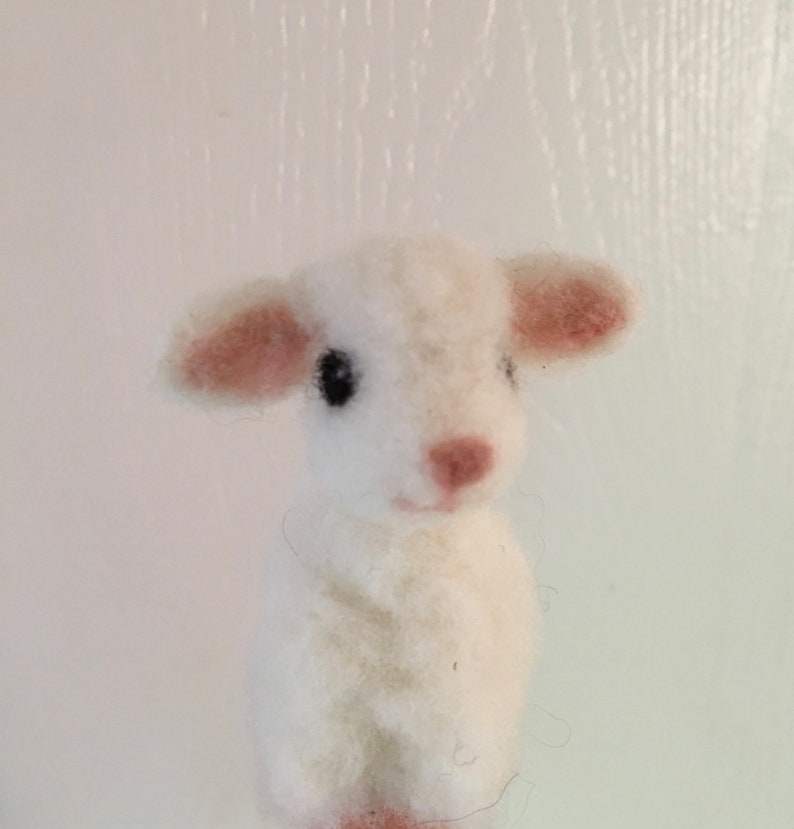 Pode incluir: Uma figura de cordeiro de feltro branco artesanal com uma textura fofa. O cordeiro tem orelhas pequenas, rosadas e um nariz a condizer. Seus olhos s&atilde;o pretos, e o design &eacute; simples e charmoso.