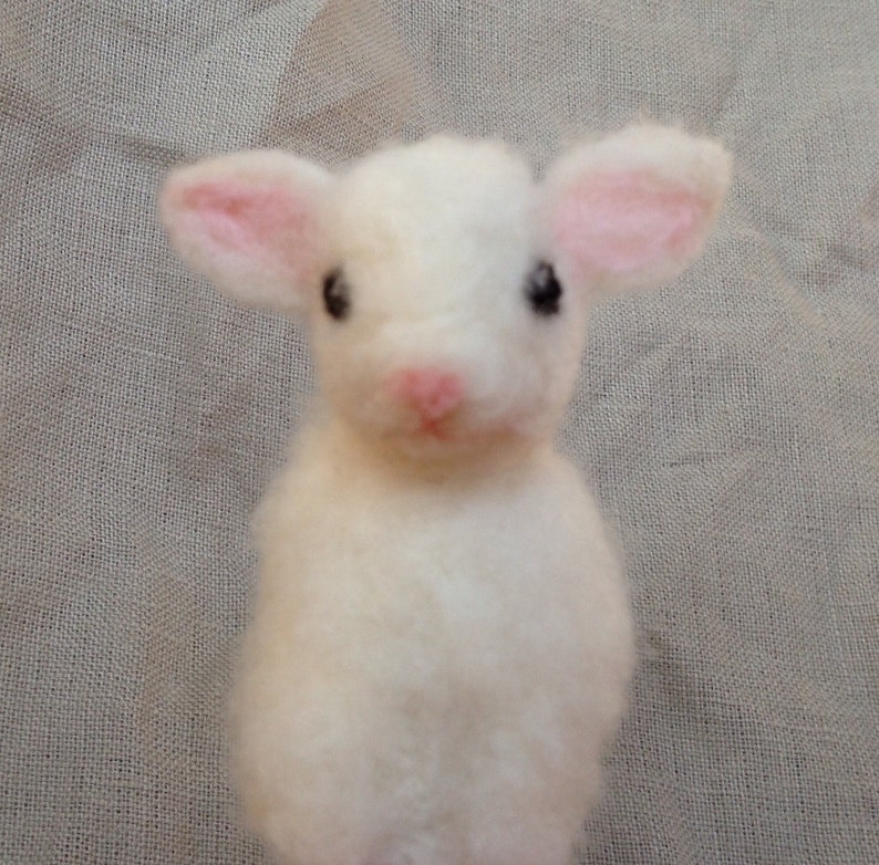 Pode incluir: Uma figura de cordeiro feita &agrave; m&atilde;o em feltro branco com o interior das orelhas e o nariz rosa. O cordeiro tem olhos pretos e uma apar&ecirc;ncia fofa e texturizada. A figura &eacute; um item artesanal charmoso.