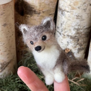 May include: A needle-felted fox figurine featuring a textured grey and brown coat with a white chest and paws. The small animal has pointed ears, black bead eyes, and a black nose, crafted with a soft, fuzzy wool finish and a detailed tail.