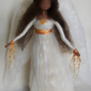 May include: A white felt angel doll with brown hair, a gold crown, and white wings. The angel is wearing a white dress with a gold sash.
