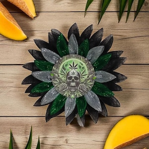 Puede incluir: Una flor decorativa con pétalos negros, verdes y plateados, con un diseño de calavera, hojas de cannabis y rosas. La flor está sobre una superficie de madera, con rodajas de mango y hojas verdes.