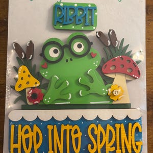 Puede incluir: Un letrero artesanal de madera con una rana verde con gafas negras, rodeada por un hongo amarillo, un hongo rojo y flores. El letrero incluye el texto "Frog Spring", "Interchangeable", "RIBBIT" y "HOP INTO SPRING" en letras amarillas.