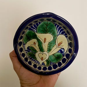 May include: A round ceramic bowl featuring a hand-painted design of white calla lilies with green leaves against a cream background. The rim is a deep cobalt blue with a repeating pattern of dark blue dots, creating a vibrant and colorful aesthetic.