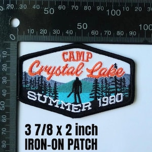 May include: Iron-on patch featuring a white background with the text "Camp Crystal Lake" in orange and black. The text "Summer 1980" is below in white. The patch is 3 7/8 inches by 2 inches.