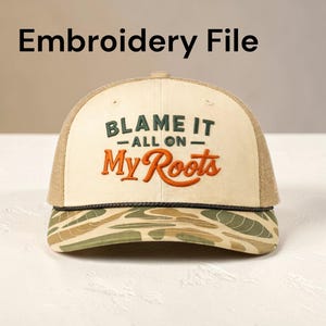 Op de afbeelding: Beige en bruine truckerpet met de tekst "BLAME IT ALL ON My Roots" geborduurd in groen en oranje. De pet heeft een camouflagepatroon op de rand en een mesh achterkant.