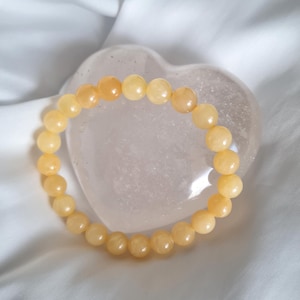 Peut inclure: Un bracelet de perles rondes de couleur jaune clair. Le bracelet est posé sur un objet transparent en forme de cœur, sur un fond blanc. Les perles ont un aspect lisse et poli.