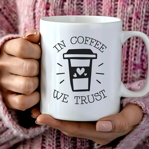 Könnte beinhalten: Weiße Keramik-Kaffeetasse mit einer schwarzen Grafik einer Kaffeetasse und dem Text "IN COFFEE WE TRUST". Die Kaffeetassengrafik enthält ein Herz. Die Tasse wird von einer Person gehalten, die einen rosa Pullover trägt.