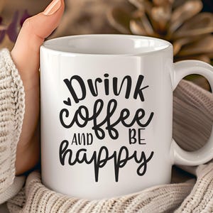 Könnte beinhalten: Weiße Keramik-Kaffeetasse mit dem Text "Drink coffee AND BE happy" in schwarzer Schrift. Die Tasse hat einen gebogenen Henkel und wird von einer Person gehalten, die einen cremefarbenen Pullover trägt. Der Hintergrund ist unscharf.