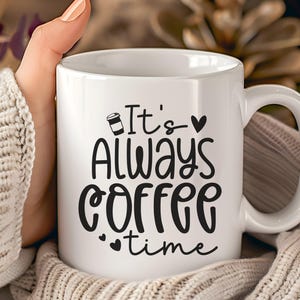 Könnte beinhalten: Weiße Keramik-Kaffeetasse mit schwarzem Aufdruck "It's Always Coffee Time". Die Tasse hat einen gebogenen Henkel und wird von einer Person gehalten, die einen cremefarbenen Pullover trägt. Der Hintergrund ist unscharf.