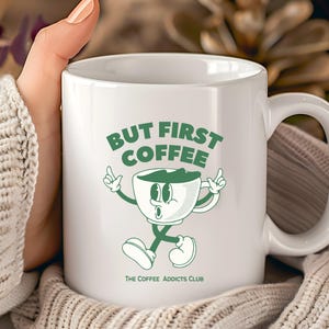 Könnte beinhalten: Weiße Keramik-Kaffeetasse mit einem grünen Cartoon-Kaffeetassen-Charakter und dem Text "BUT FIRST COFFEE". Die Tasse trägt auch den Text "THE COFFEE ADDICTS CLUB". Die Tasse wird von einer Person gehalten, die einen cremefarbenen Pullover trägt.