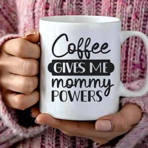 Könnte beinhalten: Eine weiße Keramik-Kaffeetasse mit dem Text "Coffee GIVES ME mommy POWERS" in schwarzer Schrift. Die Tasse hat einen großen Henkel und wird von einer Person gehalten, die einen rosa Pullover trägt.